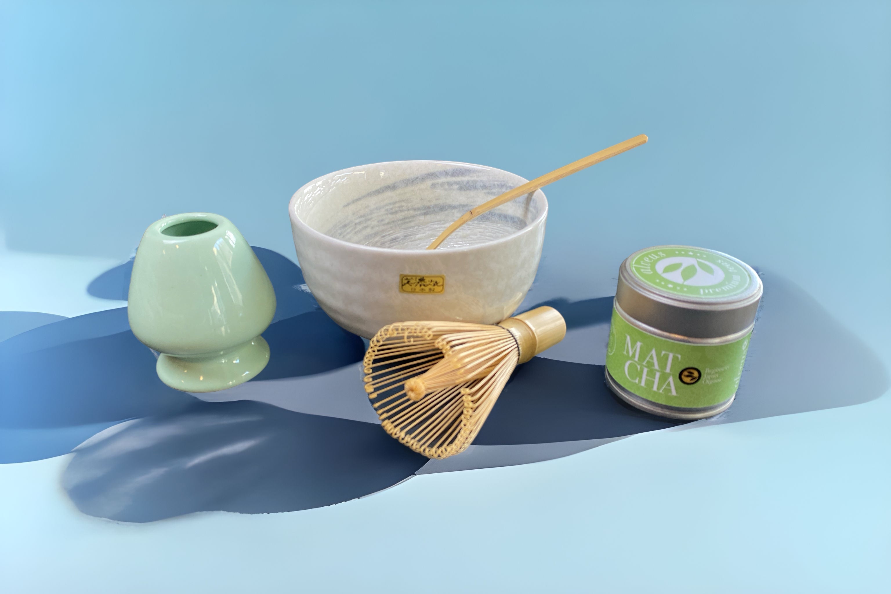 Komplettes Matcha-Zubehör-Set bestehend aus Matchabesen, Matchalöffel, Matchabesenhalter und Matchaschale, stilvoll arrangiert vor blauem Hintergrund. Perfekt für die traditionelle Matcha-Zeremonie.