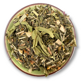 Eine Teeschale mit dem Trockenprodukt des Mango Mojito Bio, bestehend aus grünem Tee, Mangostücken, Pfefferminzblättern und Zitrusschalen, arrangiert neben einer Tasse frisch aufgebrühten, hellgrünen Tees.