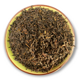 Eine Teeschale mit dem Trockenprodukt des Lapsang Souchong Bio, bräunlich-schwarze Blätter mit holziger Textur, begleitet von einer Tasse warmem, kupferfarbenem Tee.