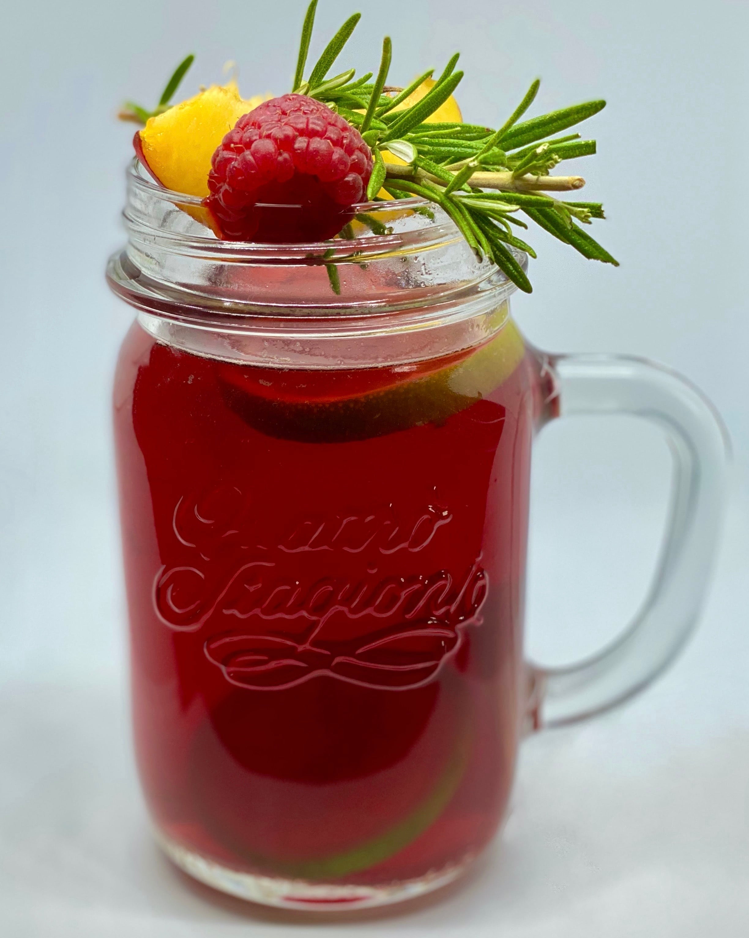 Zu sehen ist ein Eisteeglas mit dem fertigen Früchtetee und einer Dekoration aus Himbeeren, Rosmarin und Pfirsisch