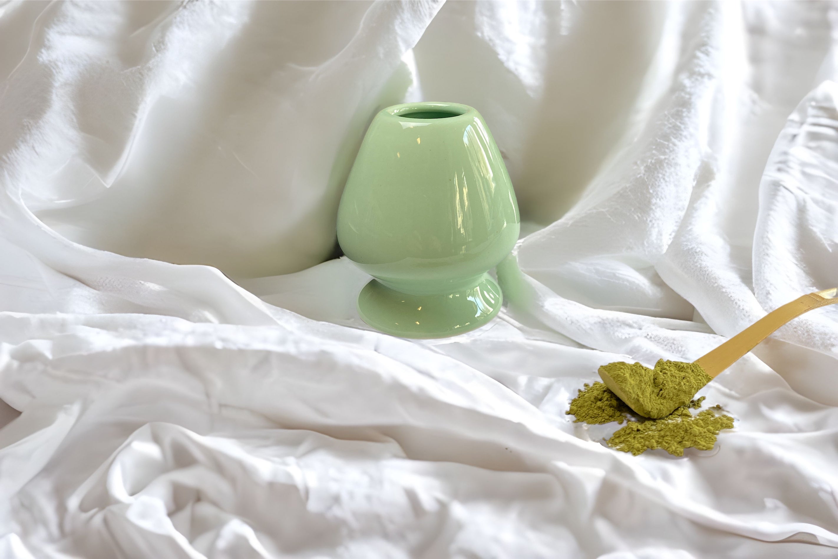 Lindgrüner Matchabesenhalter, zusammen mit einem Bambuslöffel und Matcha-Teepulver auf einem weißen Tuch arrangiert. Stilvolles Zubehör für die perfekte Matcha-Zubereitung.